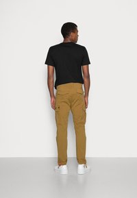 Braune Cargo-Hosen mit mehreren Seitentaschen, im geraden Schnitt und mit verstellbaren Knöchelbindungen. Kombiniert mit einem schwarzen T-Shirt und weißen Turnschuhen.