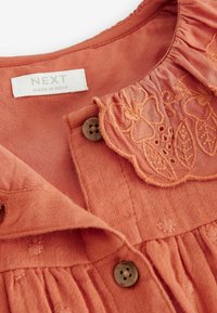 Roodbruine blouse van zachte stof met een geborduurde kraag, knoopsluiting met een houten knoop en een subtiel textuurpatroon.