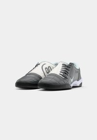 Par de sapatilhas de futsal Nike cinza e brancas com solas pretas e atacadores estampados sobre um fundo branco.