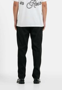 Witte t-shirt met zwarte geborduurde tekst op de achterkant, gecombineerd met zwarte broek en zwarte schoenen, gezien van de achterkant.