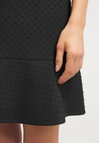 Main reposant à côté d'une jupe noire texturée avec un motif subtil de grille et de points sur un fond clair.