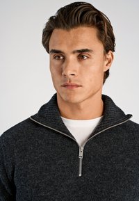 Donkergrijze trui met een hoge geribbelde kraag en een zilveren rits. De stof lijkt gestructureerd en dik, wat duidt op warmte en comfort.