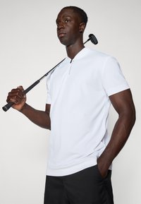 Fehér, rövid ujjú golfpóló gombos gallérral, sima textúrával és diszkrét részletekkel, fekete sorttal és golfütővel párosítva.