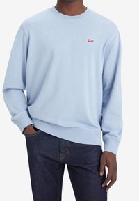 Sudadera de cuello redondo de color azul claro, hecha de suave algodón. Presenta mangas largas y un pequeño logo rojo de Levi's en el pecho. Combinada con jeans de mezclilla oscura.