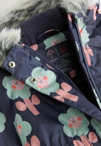 Waterproof jacket interior with floral pattern in green and pink, soft navy fleece lining, silver hardware, labels showing features.