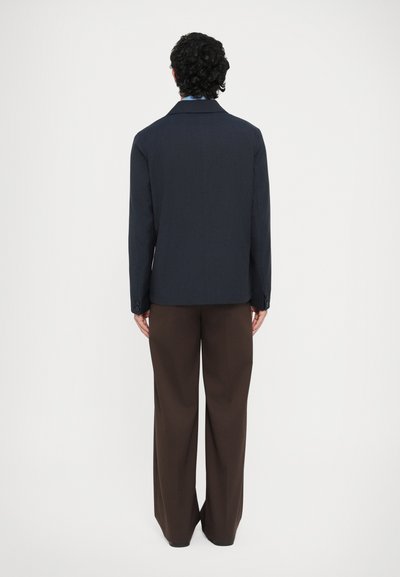 Tengerészkék zakó bevágott hajtókákkal és két gombbal, barna, széles szárú nadrággal párosítva. Sima textúrájú, szabott fazon.