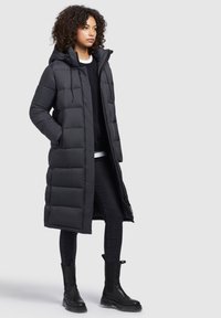 Long black puffer coat with a hood, featuring a quilted design and zipper closure. Paired with black jeans and black ankle boots.