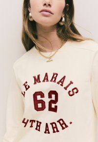 Sweatshirt crem cu text brodat roșu "LE MARAIS 62 4TH ARR." Colier cu lanț aurit și pandantiv bar, împreună cu cercei mari, texturati, în formă de cerc.