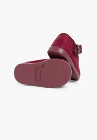 Zapatos de ante burdeos con punta redondeada, suela de goma plana y un detalle de hebilla lateral. La textura es suave, con un acabado liso.