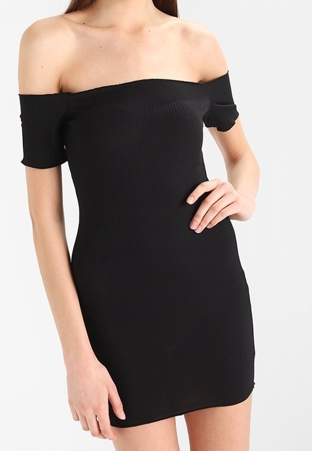 Femme portant une robe noire ajustée, épaules dénudées, manches courtes, posant avec une main sur la hanche sur un fond blanc.