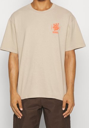 Mann trägt beige kurzärmeliges T-Shirt mit kleinem orangem Motiv und Schriftzug auf der linken Brust, kombiniert mit dunkelbraunen Hosen.