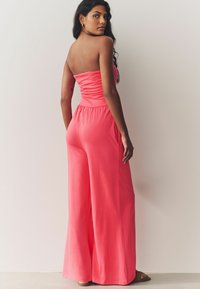 Mono sin mangas de coral con bodice recogido, piernas anchas, bolsillos laterales y textura de tela suave. Modelo mostrado desde la espalda.