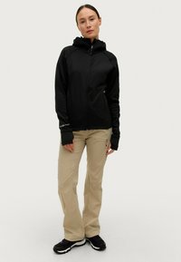 Giacca nera con cappuccio e zip frontale, abbinata a pantaloni beige chiaro. Entrambi i capi presentano un design aderente e un tessuto elastico.