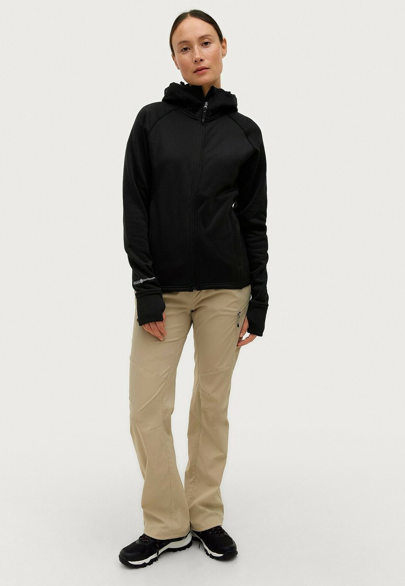 Giacca nera con cappuccio e zip frontale, abbinata a pantaloni beige chiaro. Entrambi i capi presentano un design aderente e un tessuto elastico.