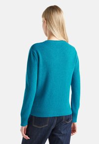 Donna con capelli biondi lisci indossa un maglione di lana teal e jeans blu scuro, vista di spalle su uno sfondo bianco.