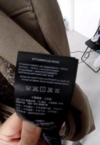 Etichetta nera con istruzioni di cura e loghi, attaccata a un tessuto beige con una texture liscia e un aspetto sartoriale.