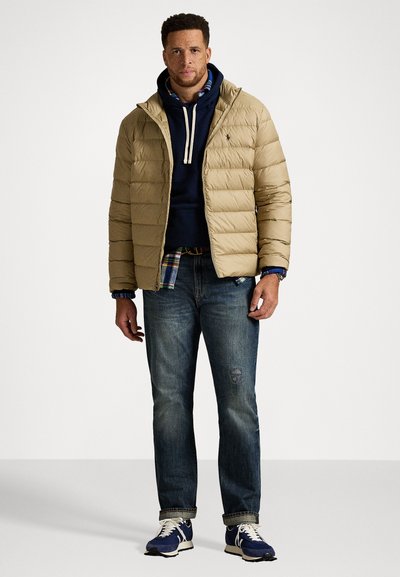 Mand iført en beige puffjakke over en marineblå hættetrøje og ternet skjorte, blå jeans og blå sneakers på en ensfarvet hvid baggrund.