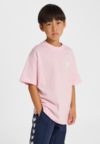 Rosa bomullst-shirt med rund hals och korta ärmar, med en liten vit logotyp på bröstet. Bärs med mörkblå byxor.