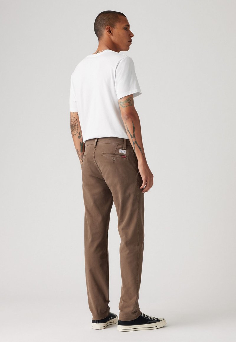 Pantalons en coton marron avec une coupe droite, des détails sur la poche arrière et une étiquette, associés à un t-shirt blanc et des baskets noires.