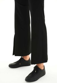 Sneaker in pelle nera con design stringato, caratterizzati da minimalisti accenti dorati e pannelli testurizzati, abbinati a pantaloni larghi neri.