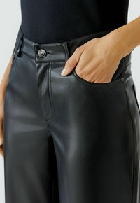 Person trägt schwarze Kunstlederhosen, mit einer Hand teilweise in der vorderen rechten Tasche, sichtbar sind der silberne Knopf und die Nähdetails.