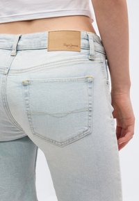 Lyseblå jeans båret af en person, der viser baglommen og en beige lædermærke på linningen.
