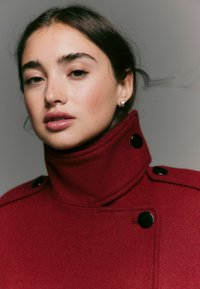 Red coat with a high collar, texture resembling wool, featuring black buttons along the front and shoulder accents.