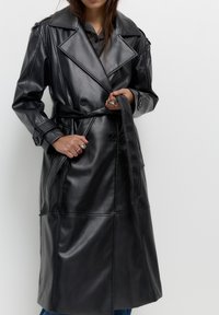 Personne ajustant une ceinture sur un long trench-coat en cuir noir avec de larges revers, portant des bagues en argent avec des pierres rouges et transparentes.