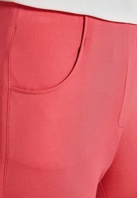 Korallenfarbene Hose mit glatter Textur, die einen abgerundeten Taschenumriss und dezente Nähdetails an der Taille und an den Beinen aufweist.