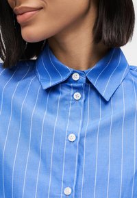 Blau gestreiftes Hemd mit einem Kragen zum Zuknöpfen, ausgestattet mit kleinen weißen Knöpfen und einer glatten Textur. Fokus auf Kragen- und Knopfdetails.