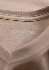 Světle béžové bavlněné tričko s kulatým výstřihem. Štítek uvádí "ombre," "100% čistá bavlna," a velikost "M." Měkká, hladká textura.