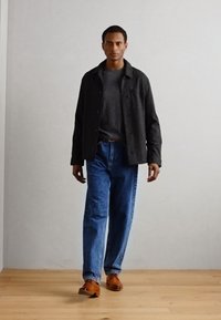 Giacca nera a bottoni sopra un maglione grigio, abbinata a jeans denim blu e scarpe marroni. Il modello sta in piedi su un pavimento di legno contro una parete chiara.