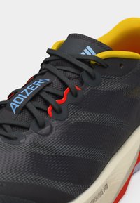 Κοντινό πλάνο μαύρου παπουτσιού τρεξίματος Adidas Adizero με κίτρινη εσωτερική επένδυση, κόκκινα κορδόνια και λευκή σόλα με τεχνολογία Lightstrike Pro.