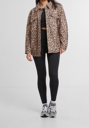 Leopardí vzorová bunda na knoflíky s dvěma náprsními kapsami, nošená přes černý crop top a černé legíny, doplněná šedými sportovními botami.