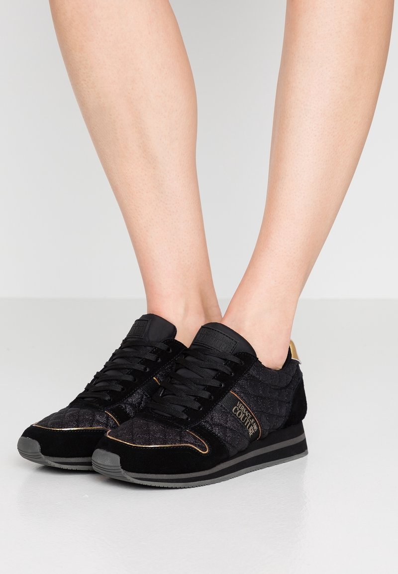 Baskets noires en daim avec texture matelassée, ornées d'accents dorés, de lacets plats et d'une semelle en caoutchouc grise.