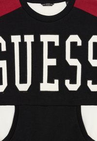 Svart hettegenser med røde skuldre, store hvite "GUESS"-bokstaver over brystet, og en forlomme i svart og hvitt.