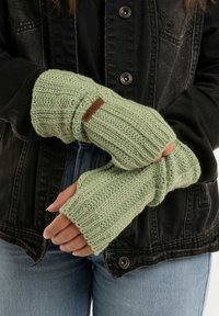 Lichtgroene gebreide vingerloze handschoenen met een gestructureerd patroon en een bruin lederen logo patch, gedragen met een zwarte jas.