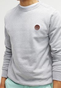 Mann trägt einen hellgrauen Rundhals-Sweatshirt mit einem kleinen braunen runden Logo auf der Brust und hellblauen Hosen.