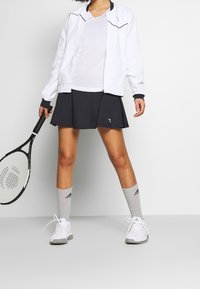 Osoba v bílé bundě, bílé košili, černé tenisové sukni, šedých ponožkách Adidas, bílých teniskách, držící černou tenisovou raketu.