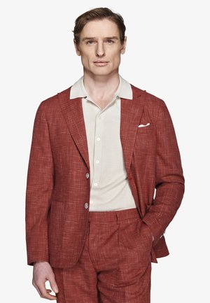 Uomo con abito testurizzato color ruggine e camicia bianca, in piedi con una mano in tasca davanti a uno sfondo semplice.