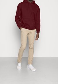 Felpa con cappuccio bordeaux e tasca frontale, pantaloni beige aderenti e sneakers bianche. Il tessuto sembra morbido con una texture liscia.