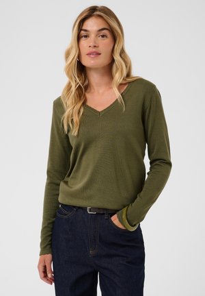 Femme aux longs cheveux blonds portant un haut manches longues vert olive à col en V et un jean bleu foncé, debout avec une main dans la poche.
