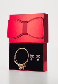 Boîte cadeau rouge avec un nœud. Contient un bracelet en or avec des charms et des boucles d'oreilles ornées de nœuds en cristal et d'accents en perles.