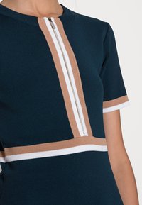 Vestido de punto en azul oscuro con mangas cortas, que presenta una cremallera central y franjas horizontales en blanco y marrón claro a lo largo del pecho.