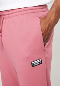 Pink sweatpants made of soft fabric, featuring an elastic waistband, side pocket, and a black Adidas logo patch on the thigh.