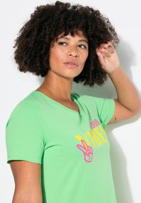Green cotton t-shirt with a V-neck, short sleeves, and a graphic print that reads "good vibes" in yellow and pink. Soft, lightweight texture.