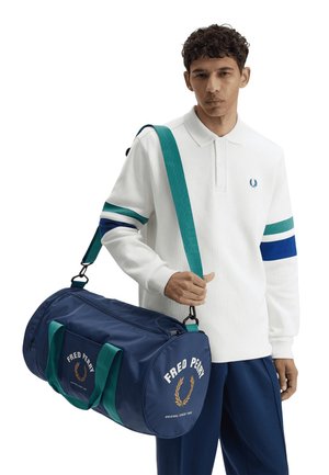 Uomo con polo bianca a maniche lunghe a righe blu-verdi porta sulla spalla una borsa da viaggio blu navy Fred Perry con manici verdi.