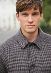 Brown checkered wool coat with a collar, featuring black buttons and a textured fabric. Model displays the coat against a blurred background.