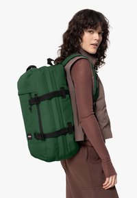 Vihreä reppu mustilla hihnoilla ja metalliosilla, jolla on rakenteellinen muoto ja sileä pinta, käytetty ruskean paidan ja liivin päällä.