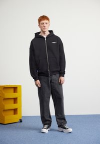 Črna zip-up kapuca s sprednjimi žepi in belim tiskom, v kombinaciji s temno sivo široko nogavičasto hlačami ter črnimi športnimi copati z belimi podplati. V bližini je rumena omara za shranjevanje.